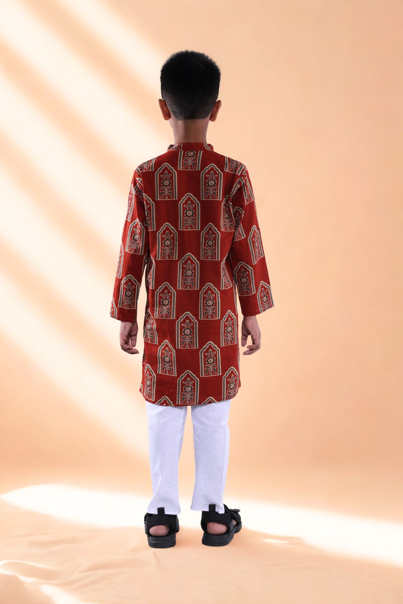 Child wearing a red patterned kurta with white pants on a beige background