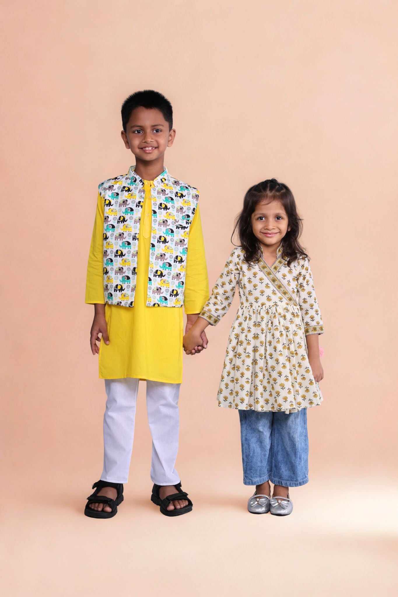 Two children holding hands, one in a yellow kurta with a patterned vest and white pants, the other in a floral dress and blue pants, on a beige background.